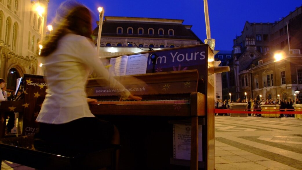 A pianist in a white jacket plays a candlelit street piano at dusk outside the Guildhall, London, with audience members visible behind a red rope barrier.