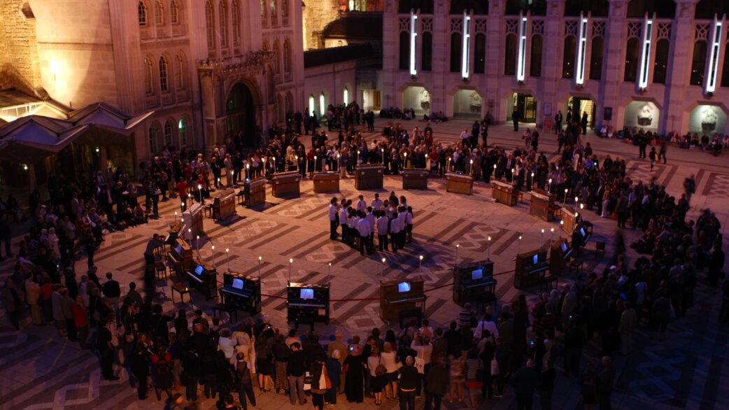 Aerial night view of 21 pianos arranged in a circle in the Guildhall courtyard, London, with performers in the centre and spectators gathered around, lit by candlelight.