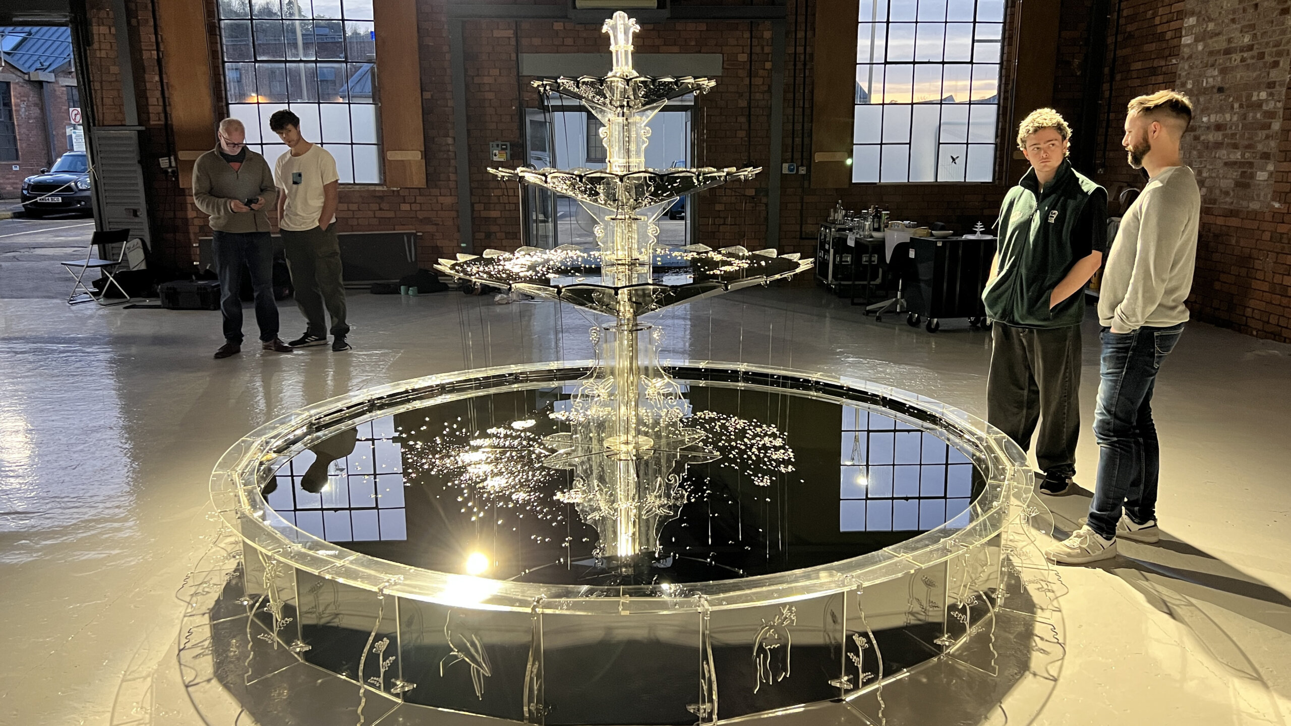 A tiered fountain sculpture approximately 2 metres tall made from clear acrylic, flowing with dark engine oil into a circular reflective pool, displayed in an industrial warehouse space with four people observing.