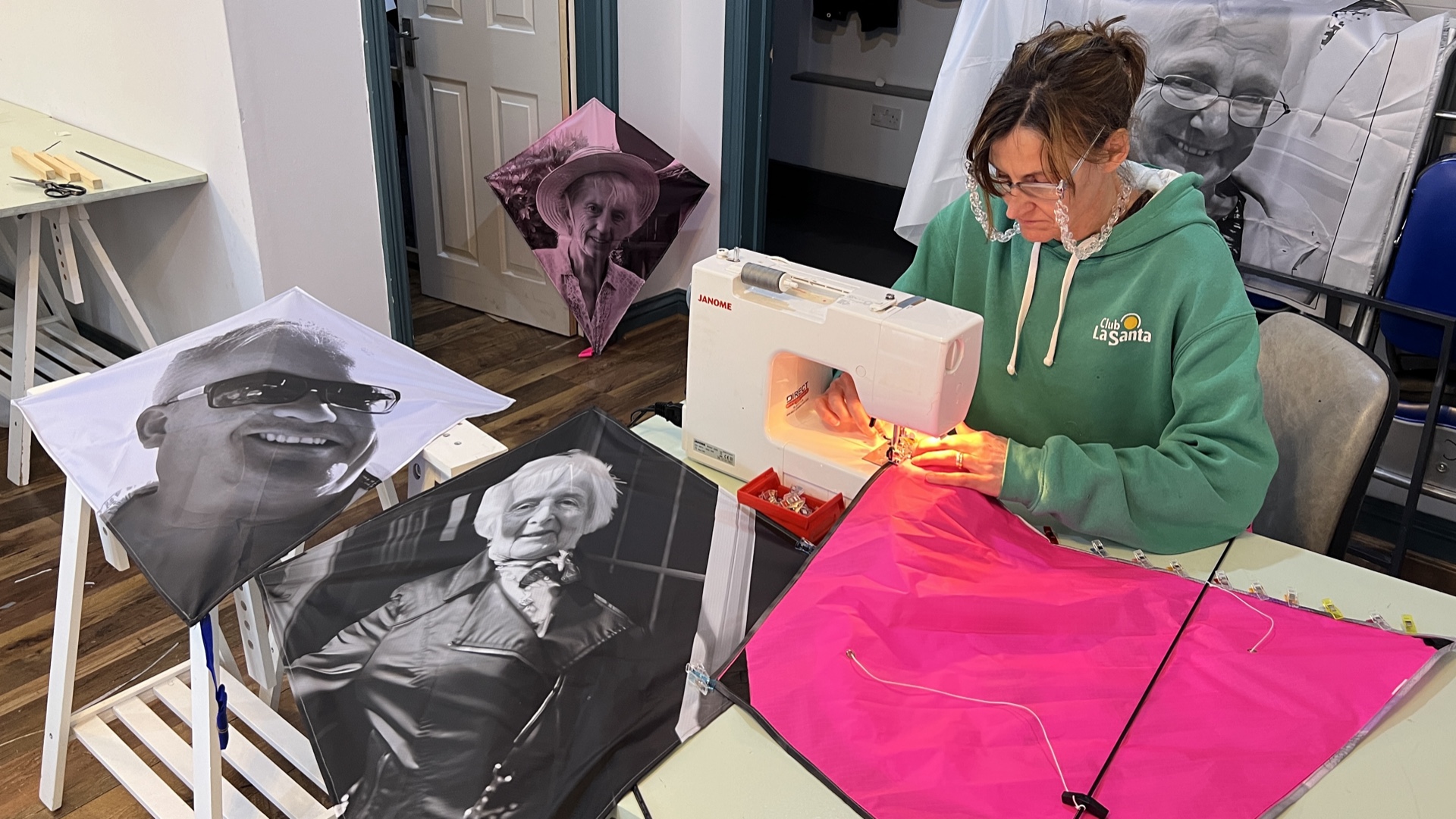 A woman in a green Club La Santa hoodie uses a Janome sewing machine to construct pink kite fabric, surrounded by completed memorial kites featuring black-and-white portrait photographs.