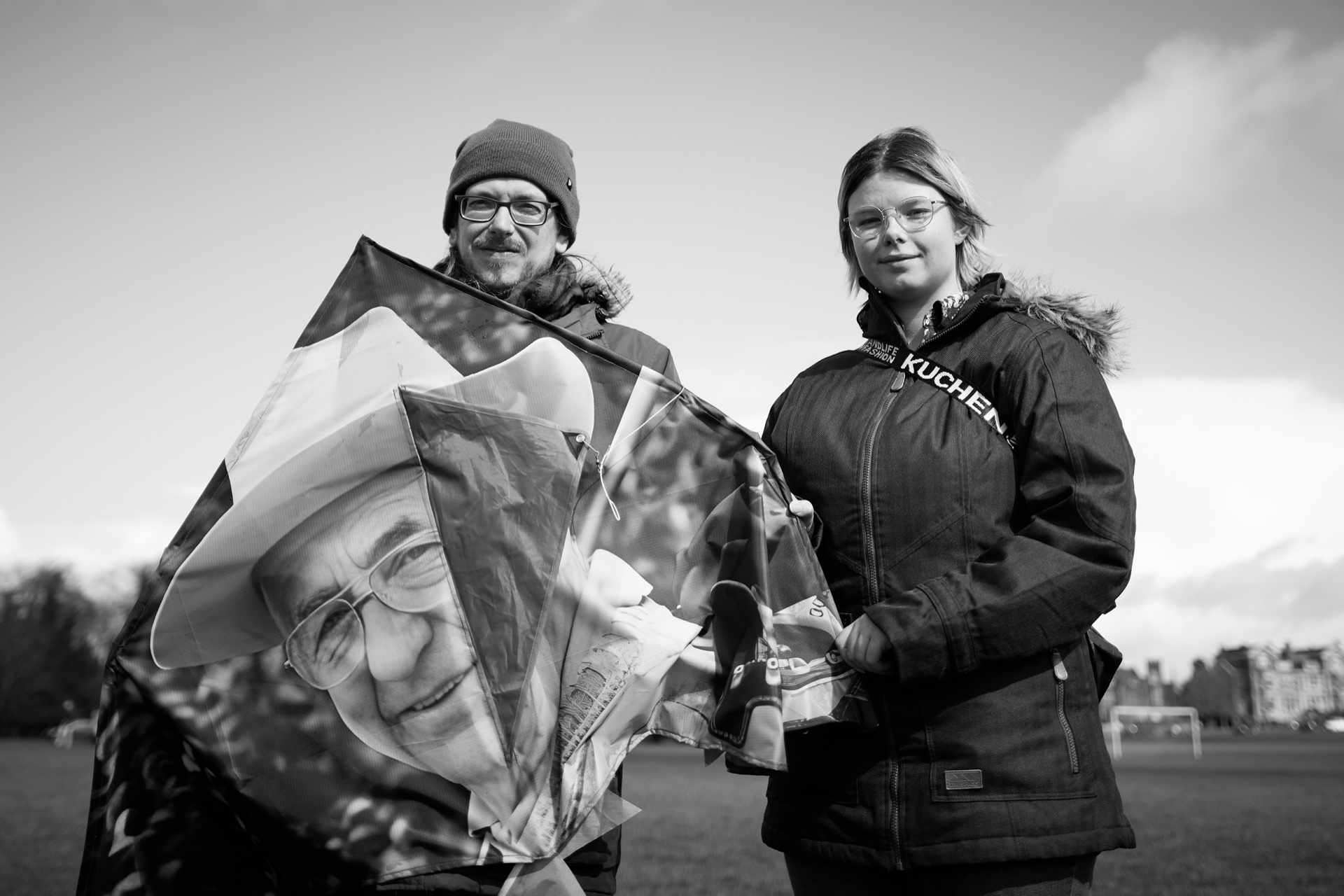Two people stand outdoors holding a kite printed with a black and white photograph of an elderly person's face, part of a Covid-19 memorial art project on Bristol Downs.