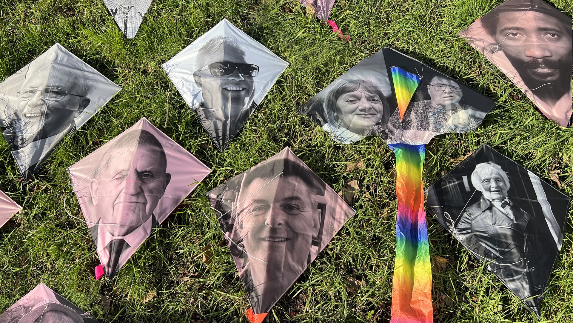 Diamond-shaped kites printed with black-and-white portrait photographs of people lie on green grass, with a rainbow-colored kite tail visible, part of a Covid-19 memorial project.