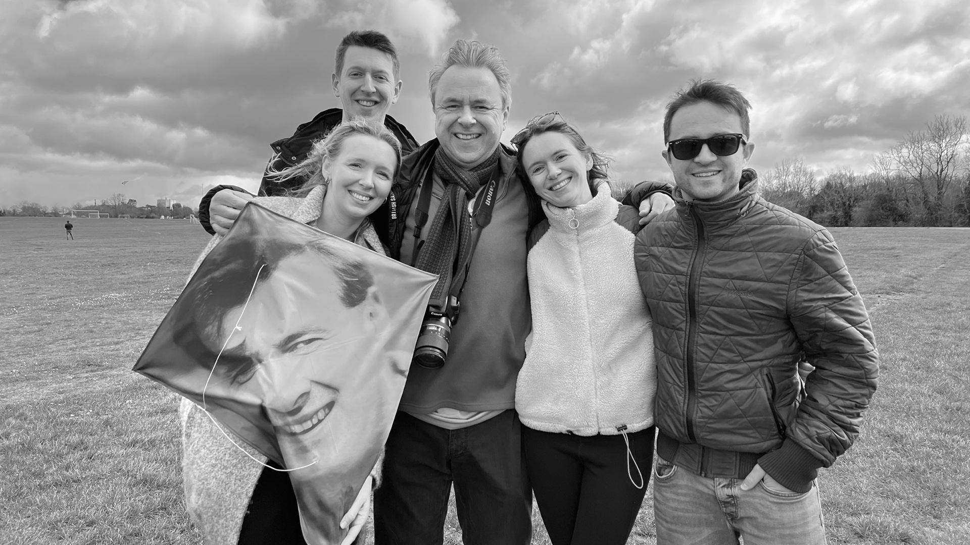 Five people pose together in a field on a cloudy day, one woman holding a kite printed with a man's smiling face. The central man wears a camera. Black and white photo.