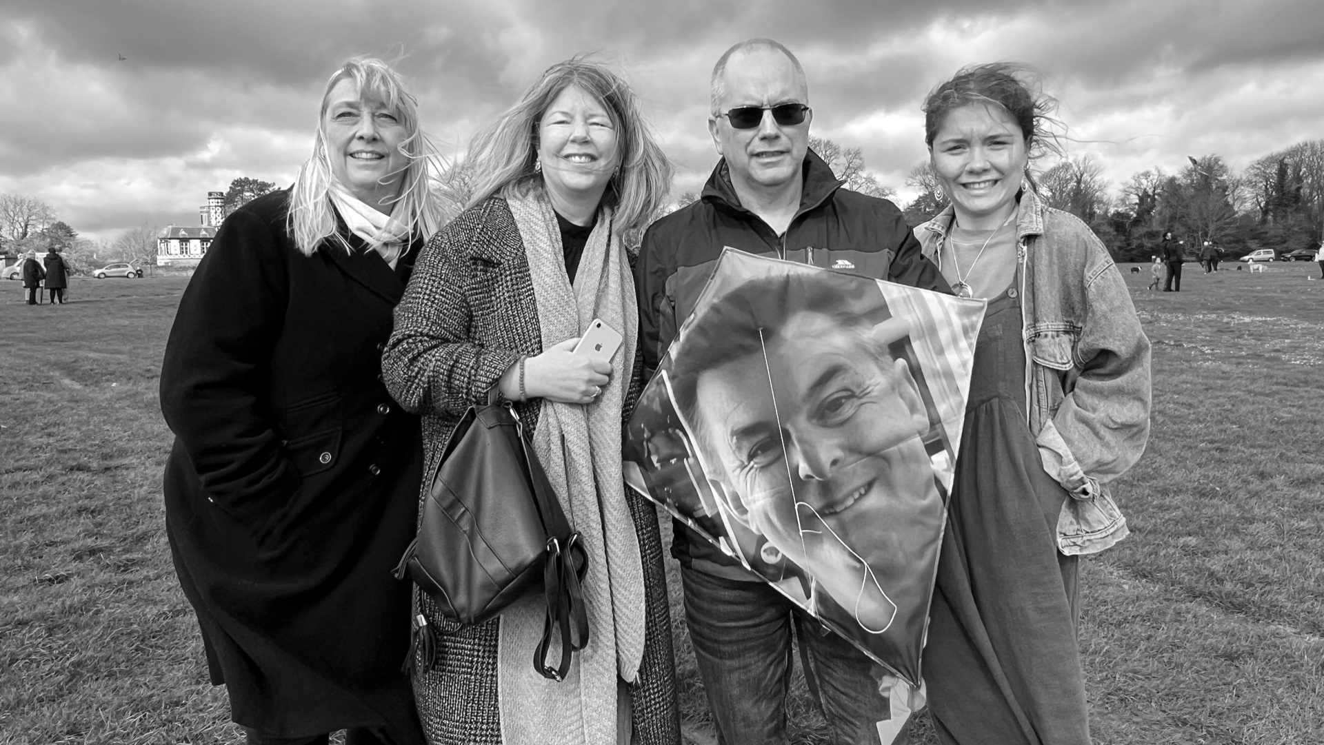 Four people stand outdoors on a windy day holding a kite printed with a smiling man's face, part of the Ascension memorial project for Covid-19 victims. Black and white photograph.