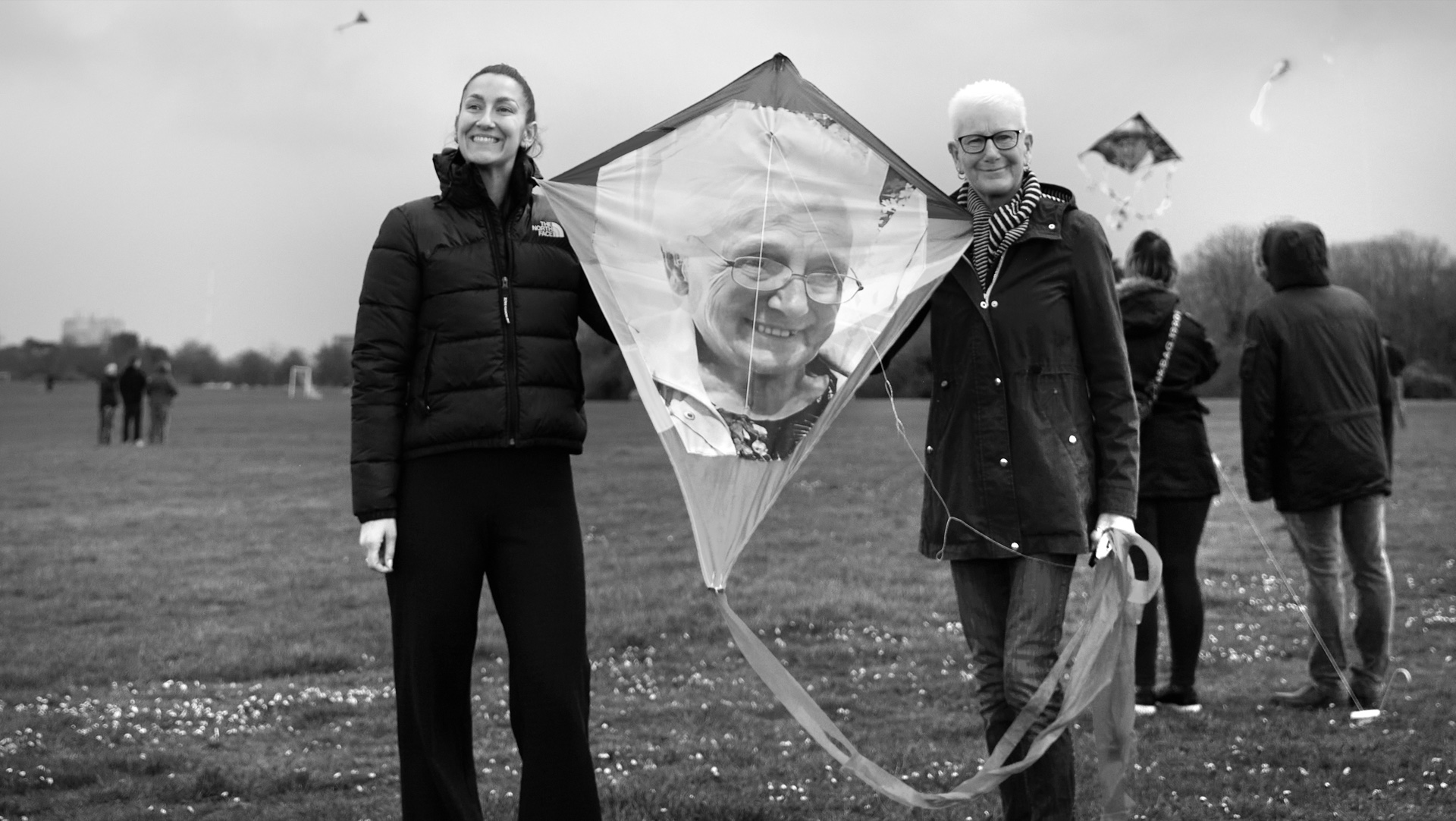 Two women stand in a grassy field holding a diamond-shaped kite featuring a printed photograph of an elderly woman with glasses. Other kite flyers visible in background. Black and white photograph.